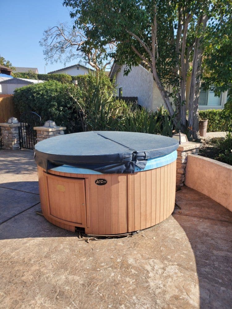 Wooden hot tub covered with a blue tarp in a backyard setting with greenery.