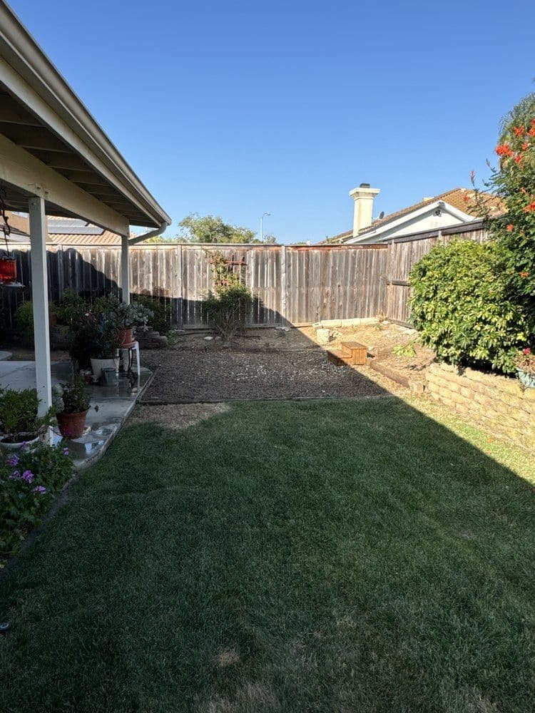 View of a backyard with a grassy area, a patio, and a wooden fence under a clear blue sky.