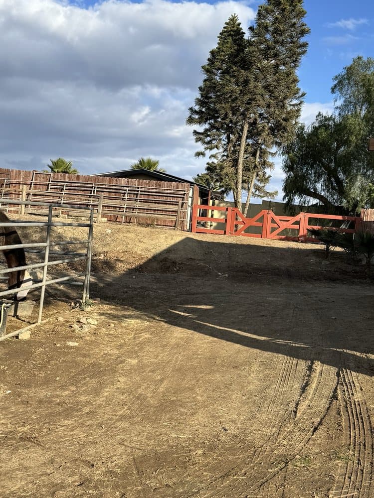 Red wooden gate and fenced area under a blue sky with trees in a rural setting.