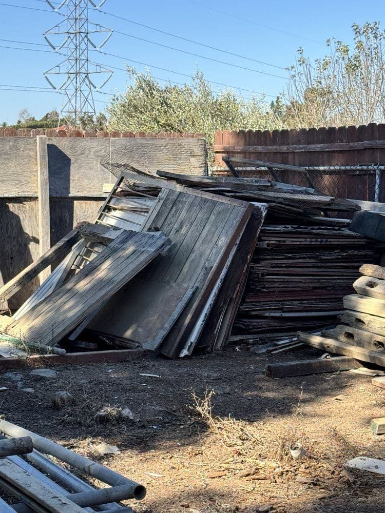 Pile of weathered wooden planks and doors in a sunny outdoor setting near a fence.