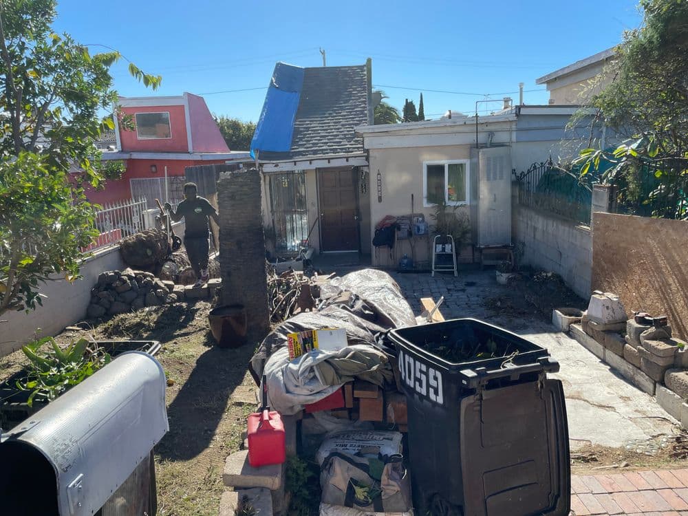 Cluttered backyard with a person cleaning near a house and trash bins under clear blue sky.