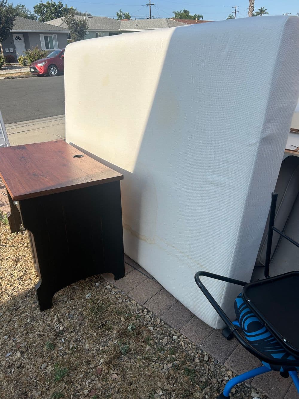 Mattress leaning against a wall next to a wooden table and a blue chair outdoors.